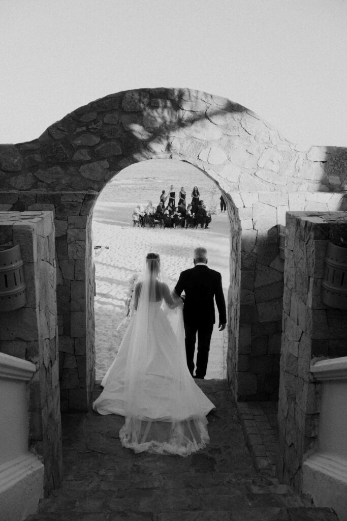 Bride with Dad walking through a stone archway before his Casa Mariposa Villa wedding ceremony